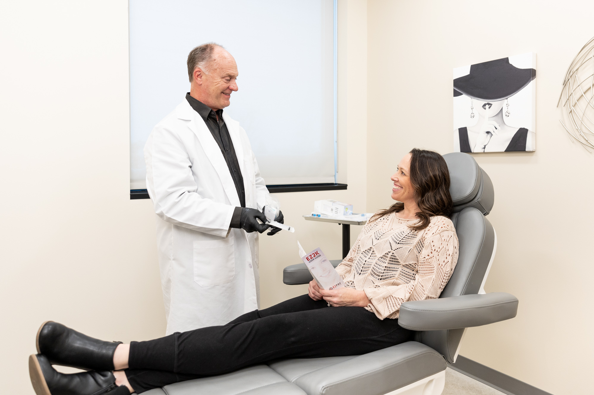 A male provider at Enrichment Skin Solutions talking to a brunette female patient sitting in a comfortable treatment chair, talking about what to expect for laser hair removal near St. Michael.