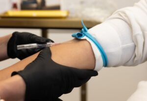 A close-up of an Enrichment Skin Solutions medical provider wearing black gloves and inserting an IV needle into a female patient's arm. IV therapy can prepare the patient's body for comprehensive body contouring in Brooklyn Park and may reduce recovery time.